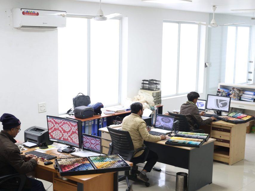 Designer creating a custom rug pattern on a digital tablet with color poms and samples nearby at Ramsha Carpets